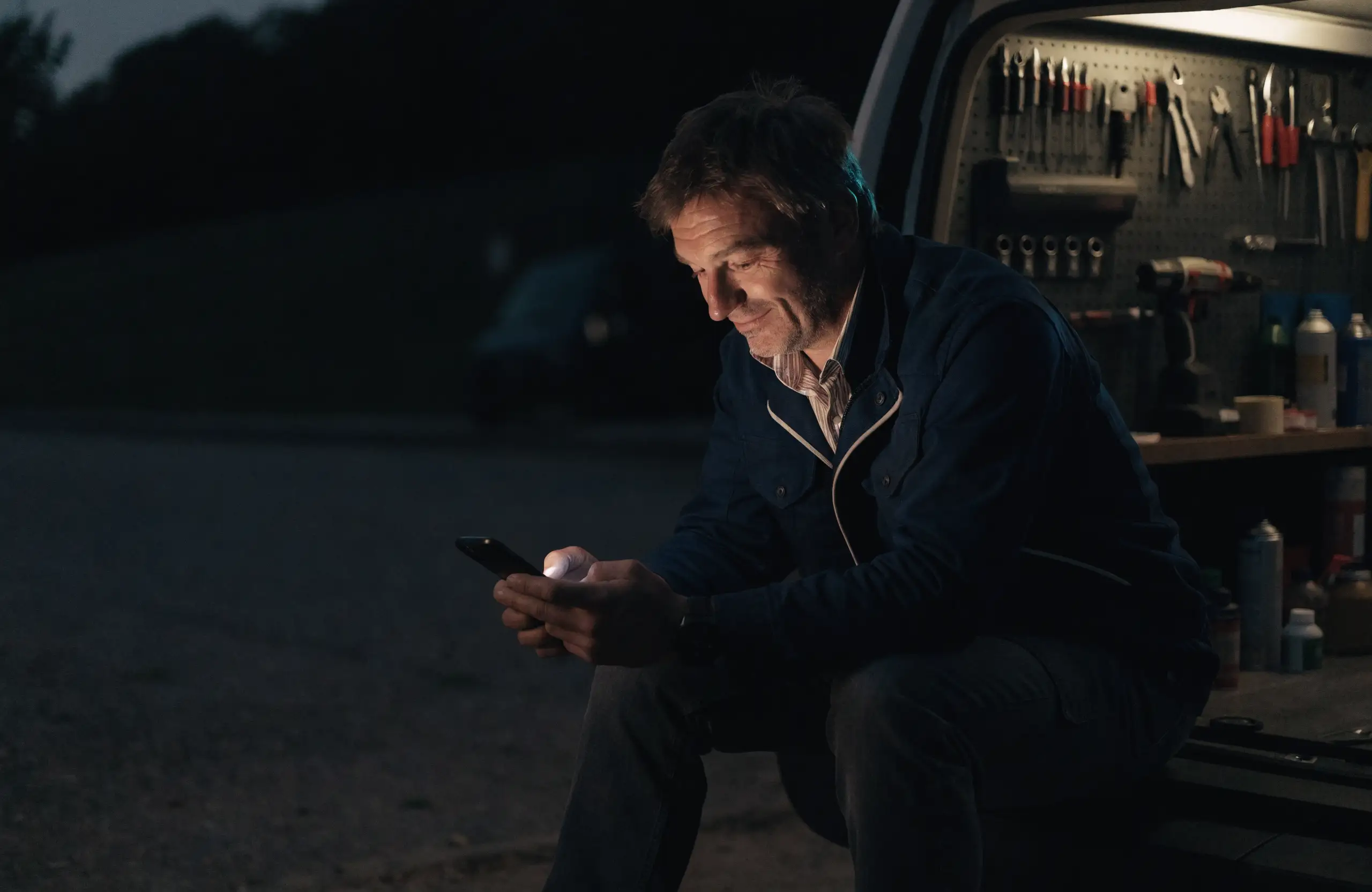 Tradesman sitting at the back of his work van at dusk, checking his phone with a smile